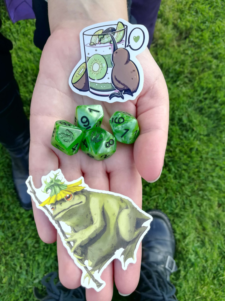 2 stickers and 3 d10 dice are seen on a hand.
The dice are mottled green.
The upper sticker consists of a glass of kiwi juice and kiwi slices. Next to it are a half kiwi and the animal kiwi, as well as a speech bubble with a green heart.
The lower sticker shows a neutral to grumpy looking frog with a dandelion hat and a rod in it's hands.
Schoes and grass can be seen in the background.